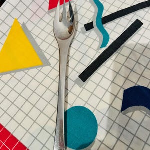 以下が含まれることがあります： 3本歯の光沢のある銀色のフォークが、白い模様のある表面に置かれています。表面には格子模様があり、赤、黄、青、黒のカラフルな幾何学模様があります。フォークは台所用品です。