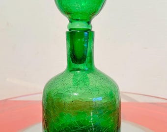 May include: A green glass decanter with a crackle finish. The decanter has a round base, a long neck, and a spherical stopper. The glass is a vibrant green color, and the crackle effect adds texture to the surface.