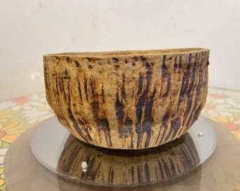 May include: A handcrafted, textured ceramic bowl with a natural, earthy aesthetic. The bowl features vertical, dark brown grooves against a light brown background. The bowl is displayed on a reflective surface.
