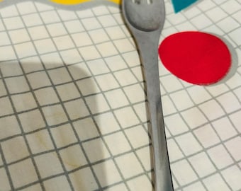 May include: A stainless steel fork with three tines, resting on a patterned tablecloth. The tablecloth features a grid pattern with yellow, teal, and red geometric shapes. The fork's handle is rectangular and tapers towards the end.