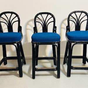 May include: Three black rattan bar stools with fan-shaped backrests and bright blue upholstered square seats. Each stool features a sturdy frame with four legs connected by a horizontal footrest bar with a metallic finish at the front.