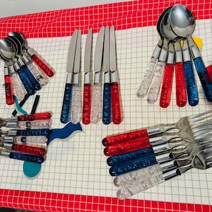 Könnte beinhalten: Eine Sammlung von Edelstahlbesteck mit transparenten, roten und blauen Acrylgriffen. Das Set umfasst Löffel, Gabeln und Messer, angeordnet auf einer rot-weiß karierten Oberfläche.