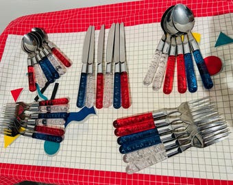 May include: A collection of stainless steel silverware with clear, red, and blue acrylic handles. The set includes spoons, forks, and knives, arranged on a red and white checkered surface.