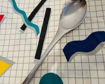 May include: A stainless steel spoon with a long handle rests on a white fabric patterned with a grid and colorful geometric shapes. The spoon is reflective, and the fabric has teal, black, and blue accents.
