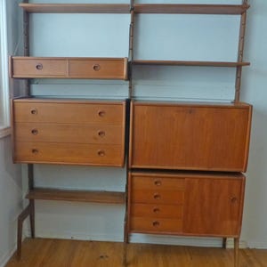 May include: A mid-century modern wall unit made of wood with shelves, drawers, and cabinets. The unit is in a light wood tone and has a simple, functional design.