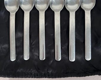 May include: Six silver spoons arranged in a row on a black surface. The spoons have a simple, classic design with oval bowls and long, slender handles. The spoons are made of a shiny, reflective metal.