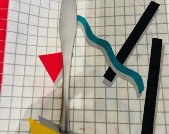 May include: A stainless steel butter knife on a white tablecloth with a grid pattern and geometric shapes in red, yellow, black, and teal. The tablecloth has a red border.