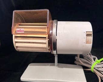 May include: Vintage Braun hair dryer with a white and tan body, mounted on a gray base. The dryer has a clear plastic cover and a dial with settings 0, 1, and 2. The Braun logo is visible.