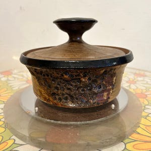 May include: A brown ceramic casserole dish with a lid. The dish has a textured, dark brown exterior with a black rim and a matching lid with a handle. The dish is displayed on a clear, round stand.