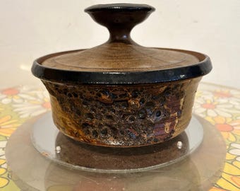 May include: A brown ceramic casserole dish with a lid. The dish has a textured, dark brown exterior with a black rim and a matching lid with a handle. The dish is displayed on a clear, round stand.