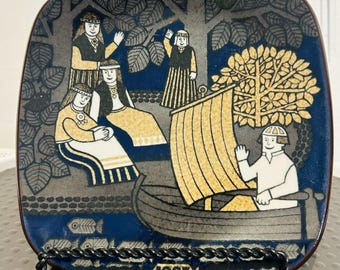 May include: A square decorative plate featuring a blue and gold illustration of four figures standing near trees and one person in a small boat with a sail. The scene includes stylized fish at the bottom and the year 1983 printed in a central rectangular box.