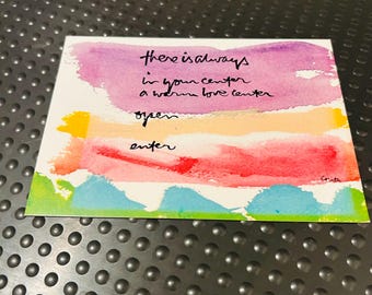 May include: A rectangular card featuring a watercolor background with horizontal bands of purple, peach, red, and blue. Black handwritten text reads: there is always in your center a warm love center open enter. The signature Corita is in the bottom right.