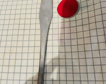 May include: A stainless steel butter knife with a long, slender handle. The knife is on a white and gray checkered cloth, with a red oval and yellow and teal geometric shapes.