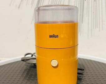 May include: A bright yellow Braun electric coffee grinder with a cylindrical body, a clear plastic lid, and a single off-white circular power button on the front. A gray power cord is visible extending from the back of the device.