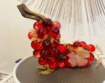 May include: A decorative cluster of translucent red glass grapes attached to a dark brown wooden stem. The grapes vary in shade from light to deep red and are accented with light tan fabric leaves and a thin, coiled green wire tendril.