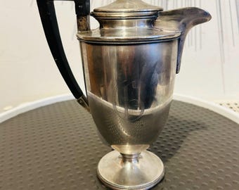 May include: A silver-toned metal pitcher with a polished finish, featuring a black angular handle, a tapered body with a circular base, and a lid topped with a small knob. The side of the pitcher is engraved with a large, stylized letter G.