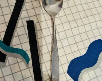 May include: A stainless steel spoon with a rounded bowl and a long, slender handle. The spoon is reflective, showing the surrounding environment. The spoon is resting on a white and gray checkered surface with blue and black geometric shapes.