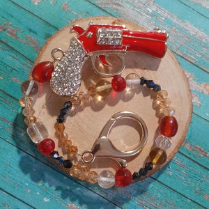 May include: A red rhinestone gun charm attached to a beaded chain with gold accents. The chain features red, gold, and black beads.