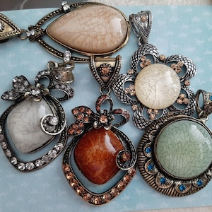 May include: A collection of ornate pendants with various shapes and colors. The pendants feature large, textured stones in shades of white, brown, and green, set in detailed metal frames with rhinestones and decorative accents.