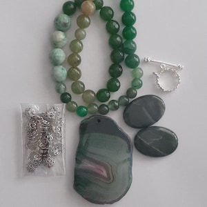 May include: An assortment of green gemstone beads, including round and faceted varieties, alongside a silver toggle clasp and decorative metal findings. Also included are two oval stones and a large, irregularly shaped pendant.