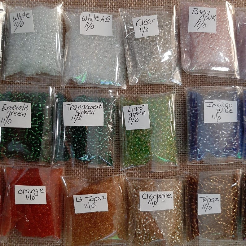 May include: A collection of small, colorful seed beads in clear plastic bags. Colors include white, white AB, clear, baby pink, emerald green, transparent green, lime green, indigo blue, orange, Lt Topaz, champagne, and topaz. Each bag has a label with the color name and size.