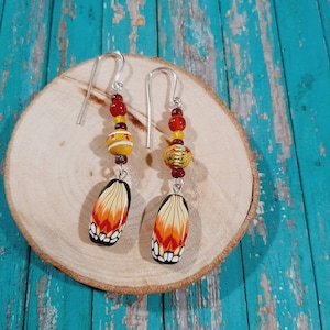 May include: A pair of dangle earrings featuring silver-tone hooks, red and yellow spherical beads, a textured yellow bead with white horizontal stripes, and a teardrop-shaped pendant with a black, orange, and white butterfly wing pattern.