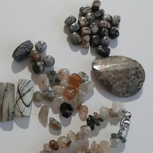 May include: Assortment of natural stone beads in various shapes and sizes, including round, oval, and rectangular. The beads are in shades of gray, white, brown, and black. Some beads have a marbled or speckled pattern. There is also a larger, irregularly shaped stone with a similar pattern.