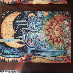 May include: A rectangular jigsaw puzzle featuring a stylized illustration of a crescent moon on the left and a sun on the right. The design includes swirling blue patterns, stars, mountains, and floral elements in vibrant orange, yellow, and blue tones.