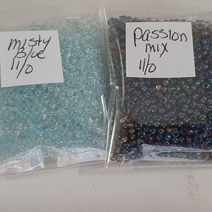 May include: Two clear plastic bags filled with small, round beads. One bag contains light blue beads labeled "misty blue 11/0." The other bag holds dark, iridescent beads labeled "Passion mix 11/0." The beads are likely for jewelry making or crafts.