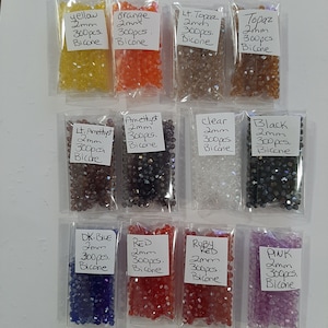 May include: Assortment of 10 small bags of bicone glass beads in various colors, including yellow, orange, light topaz, topaz, light amethyst, amethyst, clear, black, dark blue, red, ruby red, and pink. Each bag contains 300 pieces of 2mm bicone beads.