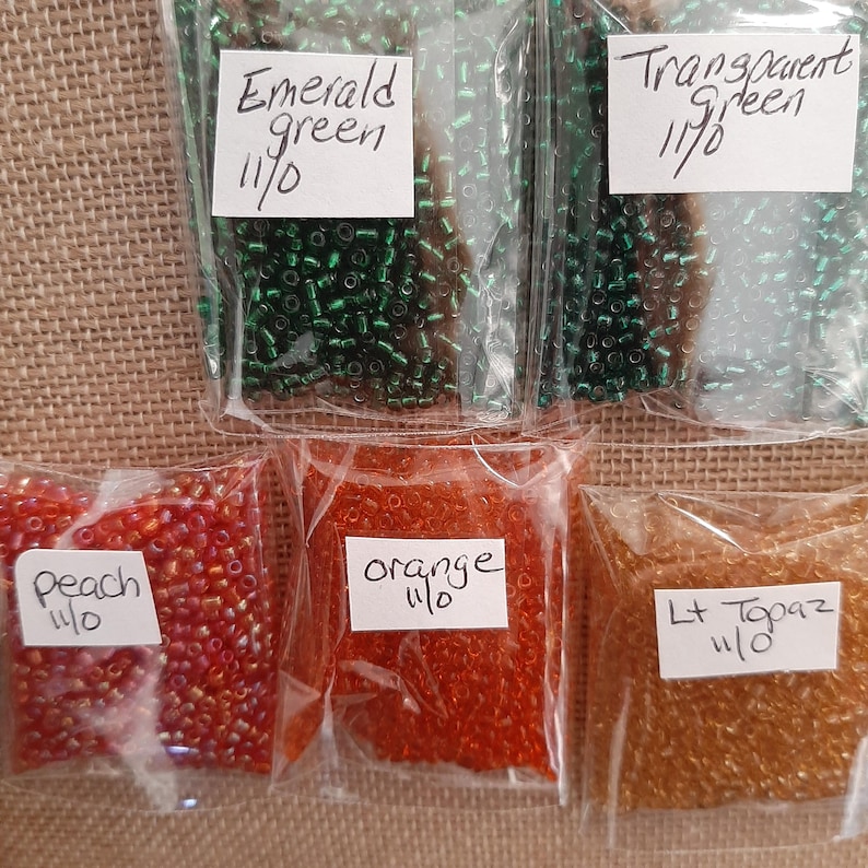 May include: Assortment of seed beads in clear plastic bags. Colors include emerald green, transparent green, peach, orange, and light topaz. Each bag has a handwritten label with the color name and size.