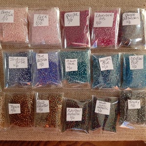 May include: A collection of small, colorful beads in clear plastic bags. The bags are labeled with color names like "Baby Pink," "Purple," and "Gold Rainbow." The beads appear to be for crafting or jewelry making.