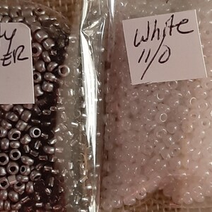 May include: Two clear plastic bags filled with small beads. One bag contains misty silver beads, and the other contains white beads. Each bag has a handwritten label with the color name and "11/0".