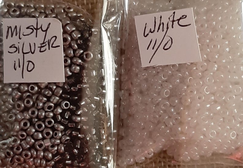 May include: Two clear plastic bags filled with small beads. One bag contains misty silver beads, and the other contains white beads. Each bag has a handwritten label with the color name and "11/0".