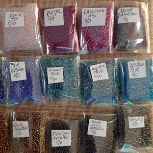 May include: A collection of small, colorful seed beads in clear plastic bags. The bags are labeled with color names like "Pink," "Purple," "Lavender AB," and "Blue Passion." The beads are in various shades, including pink, purple, blue, and metallic colors.