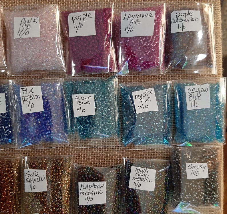 May include: A collection of small, colorful seed beads in clear plastic bags. The bags are labeled with color names like "Pink," "Purple," "Lavender AB," and "Blue Passion." The beads are in various shades, including pink, purple, blue, and metallic colors.