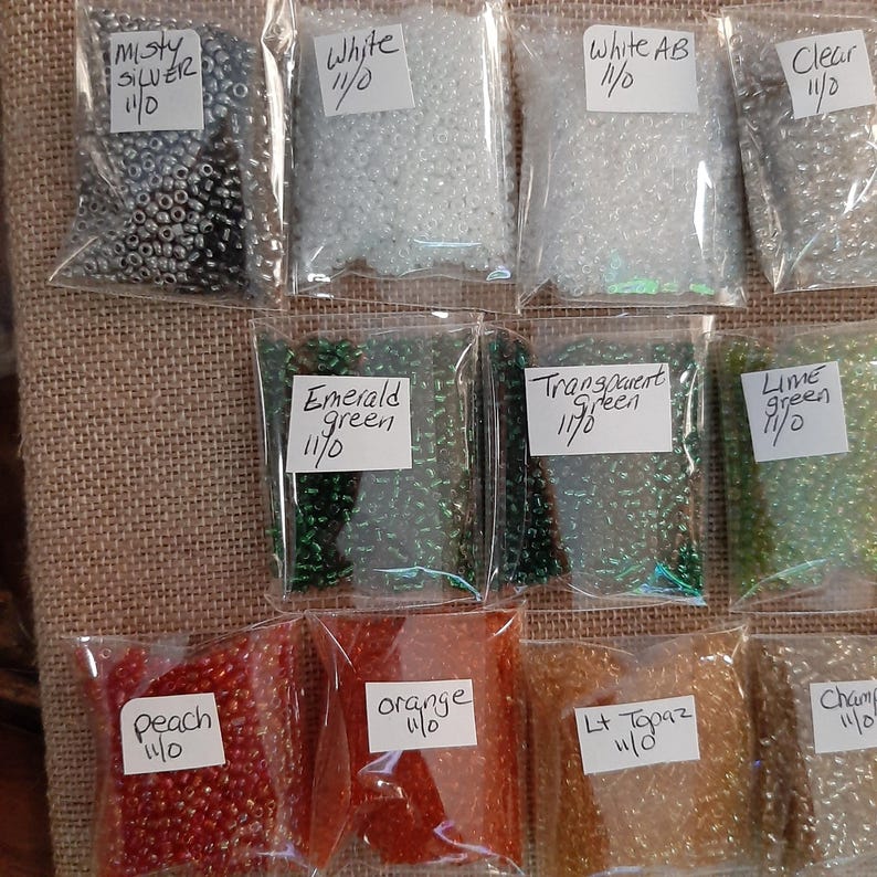 May include: A collection of small, colorful beads in clear plastic bags. Colors include silver, white, emerald green, transparent green, lime green, peach, orange, and light topaz. Each bag has a label with the color name and size.