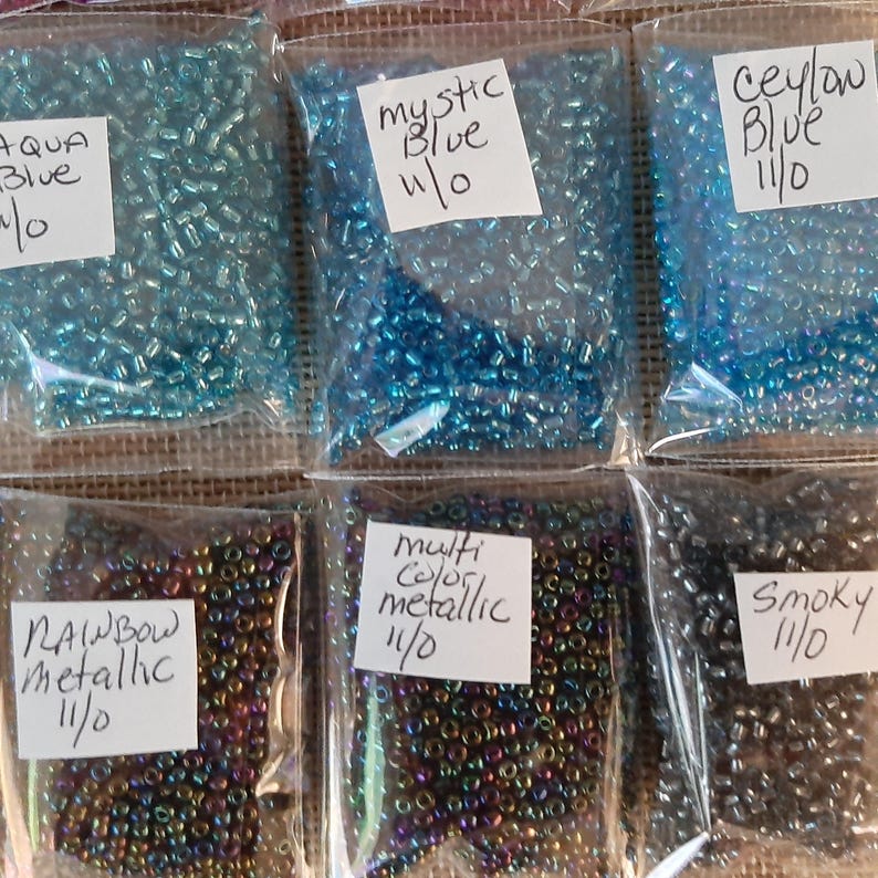 May include: Assortment of small, colorful seed beads in clear plastic bags. The bags are labeled with names like "Aqua Blue," "Mystic Blue," "Ceylon Blue," "Rainbow Metallic," "Multi Color Metallic," and "Smoky." The beads are 11/0 size.