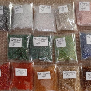 May include: A collection of small, colorful beads in clear plastic bags. Colors include silver, white, clear, pink, green, blue, orange, and gold. Each bag has a label with the color name and size.