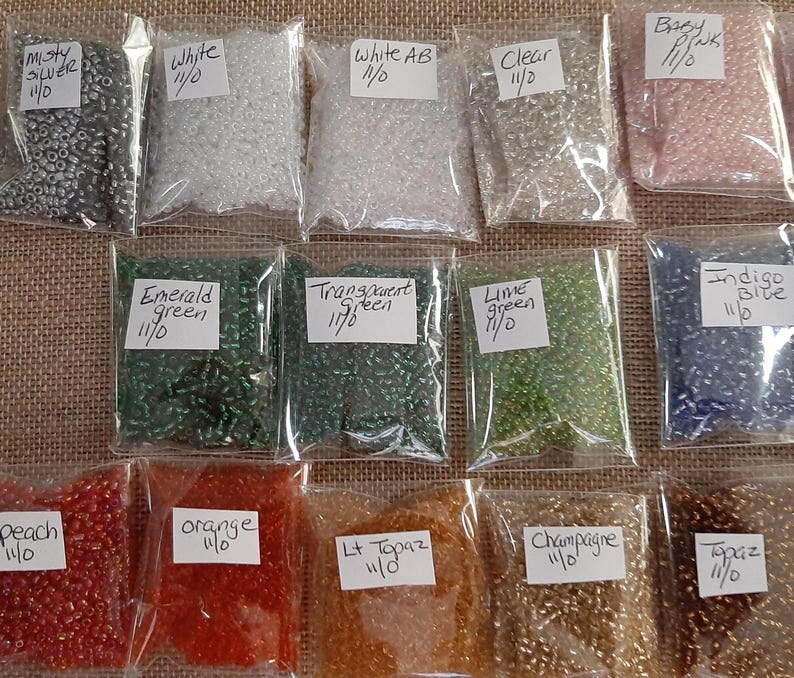 May include: A collection of small, colorful beads in clear plastic bags. Colors include silver, white, clear, pink, green, blue, orange, and gold. Each bag has a label with the color name and size.