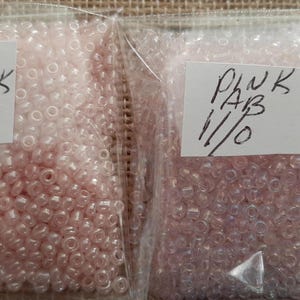 May include: Two clear plastic bags filled with small, light pink beads. One bag is labeled "BABY PINK 11/0", and the other is labeled "PINK AB 11/0". The beads appear to be seed beads, commonly used for jewelry making and crafts.