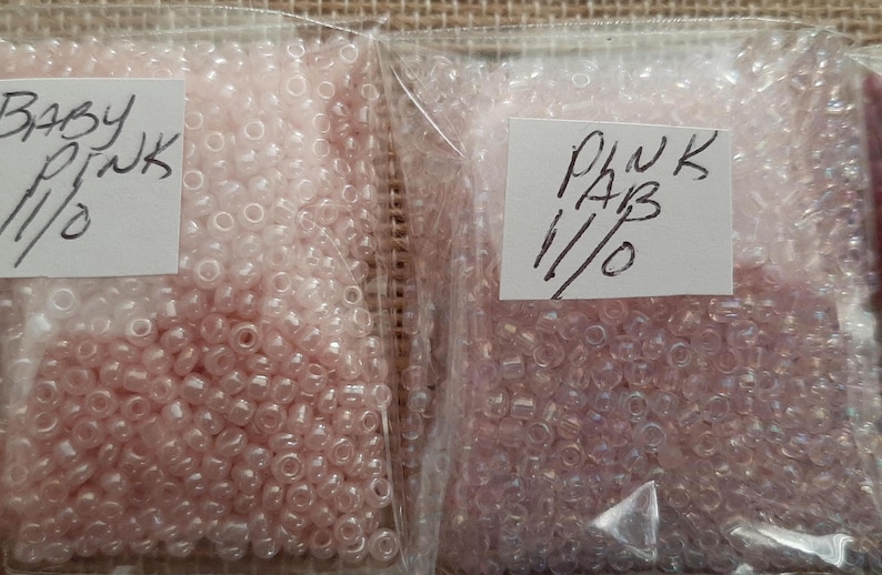 May include: Two clear plastic bags filled with small, light pink beads. One bag is labeled "BABY PINK 11/0", and the other is labeled "PINK AB 11/0". The beads appear to be seed beads, commonly used for jewelry making and crafts.