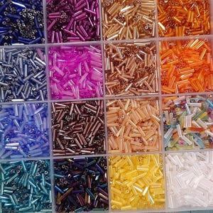 May include: A clear plastic storage container with multiple square compartments filled with an assortment of small, cylindrical glass bugle beads in a wide variety of vibrant colors including blue, pink, orange, yellow, green, white, and metallic tones.