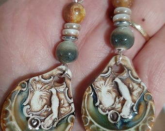 May include: Handmade earrings featuring a teardrop-shaped ceramic pendant with a bird design in white, brown, and blue. The earrings have a beaded accent with brown, white, and gray beads, and silver-tone ear wires.