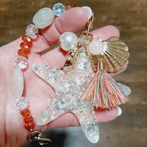 May include: A sparkly iridescent starfish charm with a colorful tassel, white and gold beads, and a pink and gold seashell charm. The charm is attached to a silver chain with a lobster clasp.