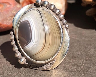 May include: A silver ring with a brown and white agate stone set in a silver bezel. The ring is decorated with small silver beads around the bezel.