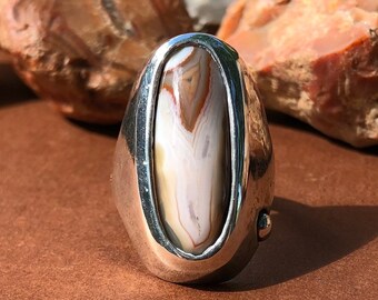 May include: A silver ring with an oval-shaped agate stone set in a bezel setting. The agate stone has brown, white, and gray swirls.