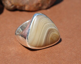 May include: A silver ring with a triangular, light brown stone featuring a striped pattern. The ring has a polished silver band and a bezel setting. The stone's color is a mix of beige and brown, with a smooth, polished surface.