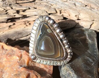 May include: A silver ring with a triangular agate stone set in a silver bezel. The stone has brown, white, and gray banding.