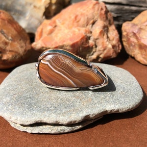 May include: A brown agate stone with white and brown banding is set in a silver ring. The ring is displayed on a gray stone.